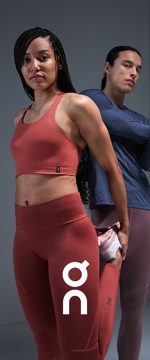 Roter Sport-BH und hoch taillierte Leggings, beide mit glatter Textur und Logo-Details. Graues Langarmshirt und helle rosa Schuhe im Hintergrund.