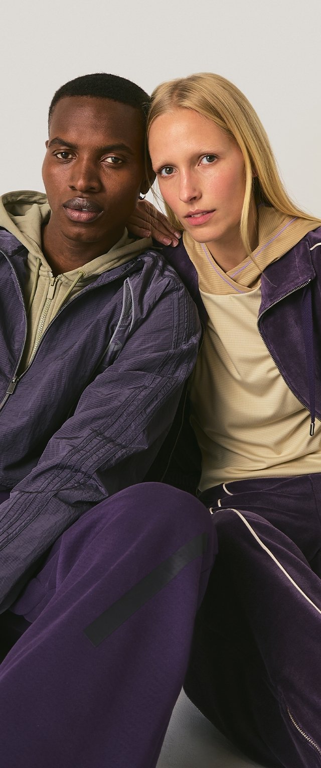 Dos personas están sentadas juntas, vistiendo chaquetas con capucha y pantalones morados a juego. Las chaquetas tienen un tejido texturizado y cremalleras, con diseños en capas.