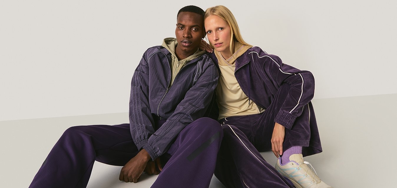 Dvě modelky sedí na podlaze a mají na sobě sladěné fialové teplákové soupravy, jedna s béžovou mikinou s kapucí a druhá s béžovým tričkem a bílými teniskami.