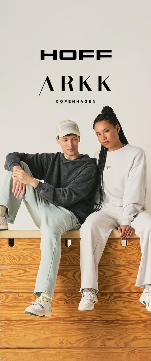 Due modelli seduti su una panchina di legno con abbigliamento casual athleisure; uomo con felpa nera con cappuccio e cappellino, donna con felpa bianca e pantaloni, con i loghi HOFF e ARKK Copenhagen sopra.