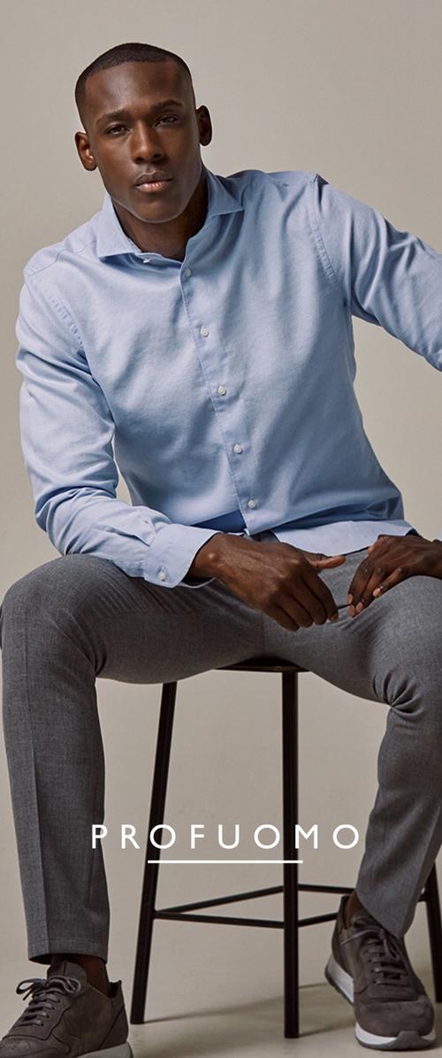 Lichtblauw overhemd van een zachte stof, gecombineerd met grijs op maat gemaakte broek. Model zit op een zwarte metalen kruk.