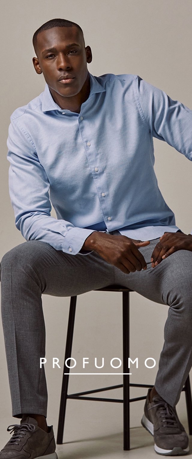 Helles blaues Hemd aus einem weichen Stoff, kombiniert mit grauen, maßgeschneiderten Hosen. Modell sitzt auf einem schwarzen Metallhocker.