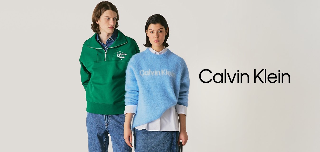 Muž v zeleném svetr s čtvrtkovým zipem a džíny stojí vedle ženy v modrém svetru Calvin Klein, džínové sukni a držící černou kabelku.