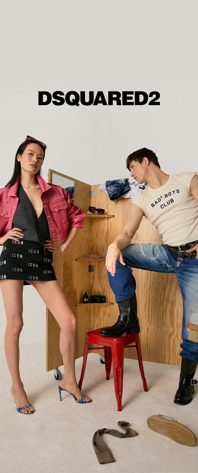Frau in pinker Jacke und schwarzem ICON-Rock posiert mit Mann in weißem "BAD BOYS CLUB" Shirt und Jeans, roter Hocker, Holzhintergrund, DSQUARED2 Logo oben.