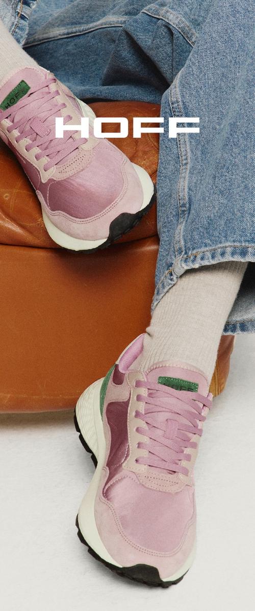 Lässige rosa und violette Sneaker mit grünen Akzenten, getragen mit cremefarbenen Socken und blauen Jeans, ruhend auf einem braunen Ledersessel.
