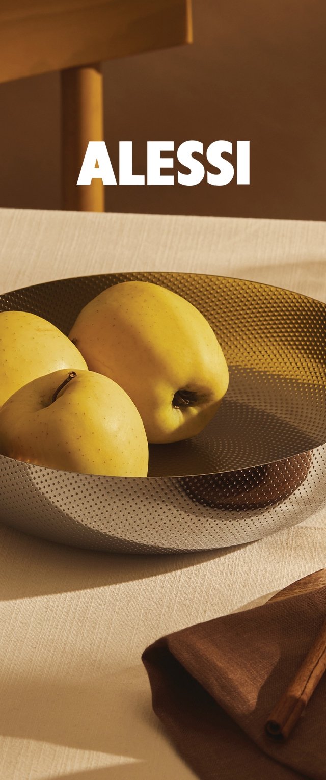 Alessi metalen kom met een geperforeerd ontwerp, waarop drie gele appels liggen, geplaatst op een lichte stoffen ondergrond met een bruine servet en een kaneelstokje.
