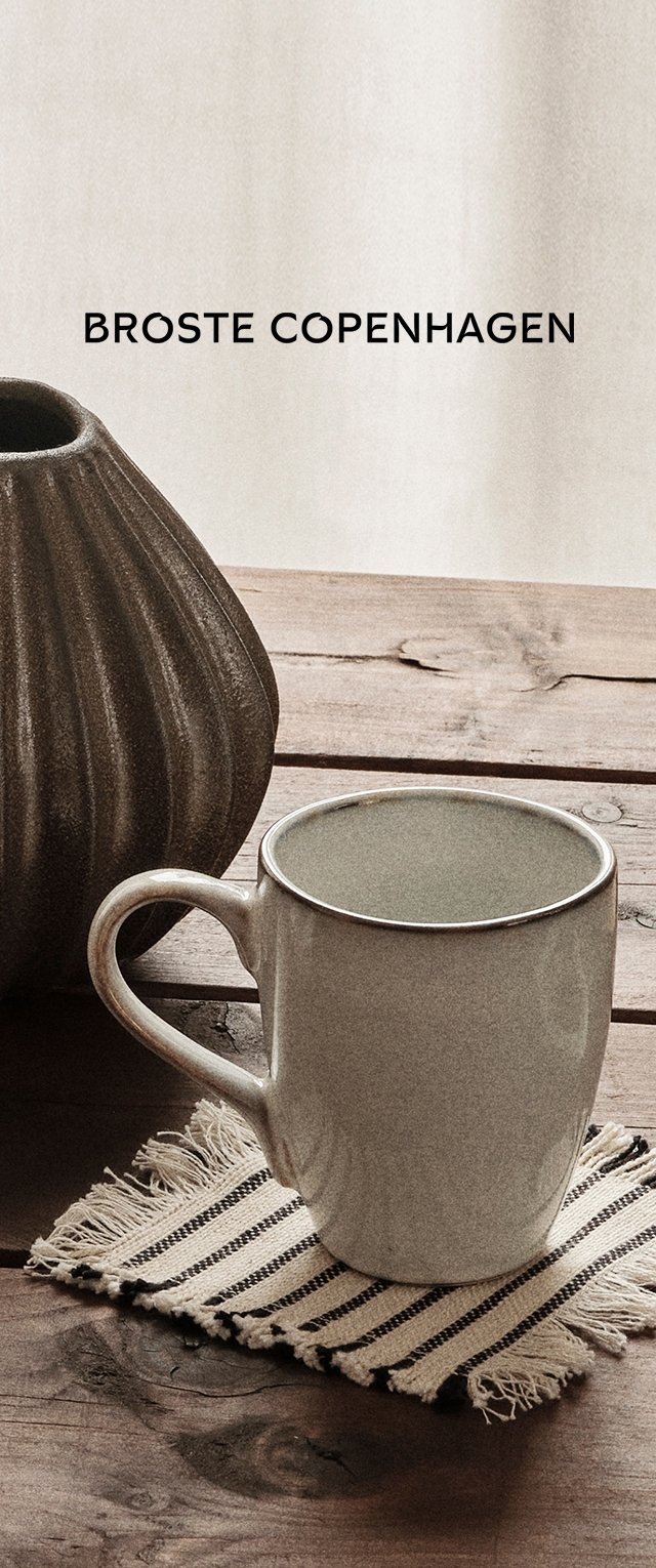 Tazza in ceramica bianca con corpo rotondo e manico, posata su un sottobicchiere intrecciato a righe, accanto a un vaso in ceramica scura e texturizzata su una superficie in legno.