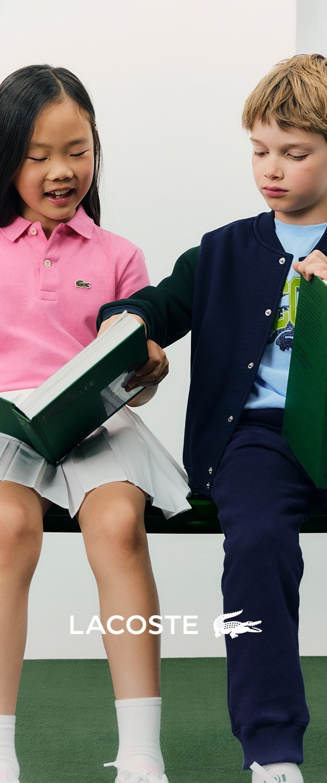 Deux enfants lisent un livre vert. La fille porte un polo rose et une jupe blanche ; le garçon a un cardigan marine et un t-shirt bleu.