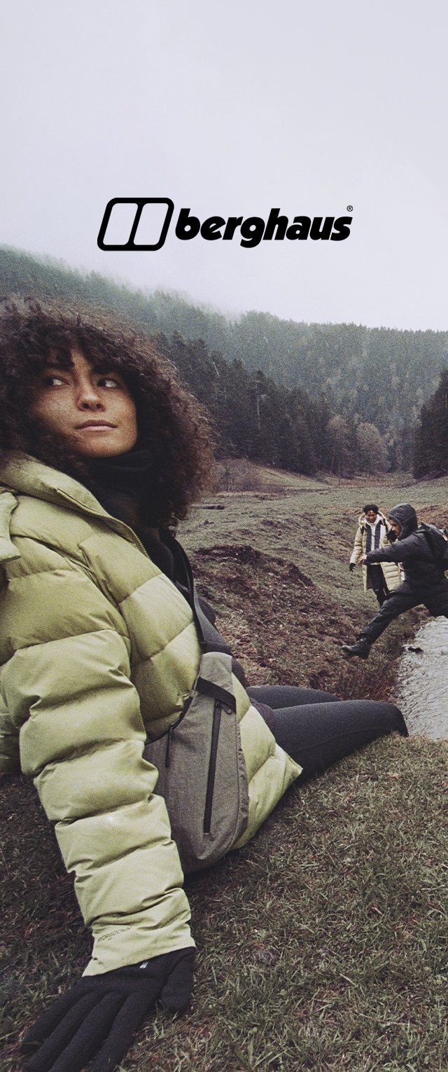 Frau in grüner Jacke und Handschuhen sitzt auf einem grasbewachsenen Hügel; zwei Personen überqueren einen kleinen Bach im bewaldeten Tal dahinter; Berghaus-Logo oben.
