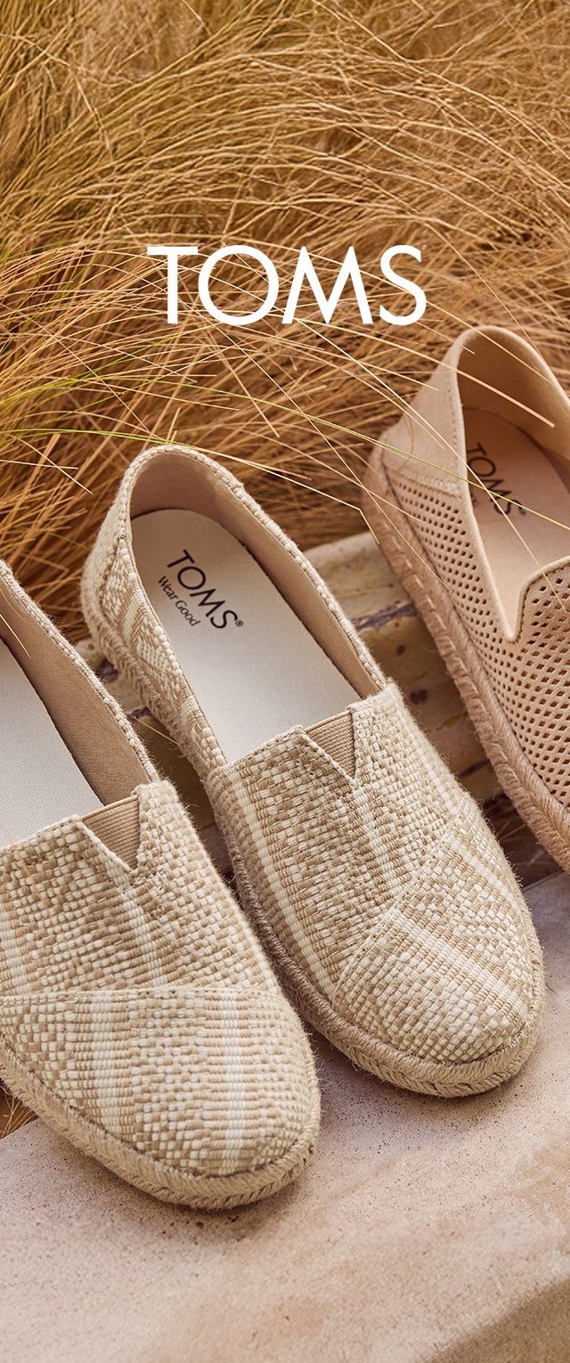 Drei Paar beige TOMS Slipper in gewebten und perforierten Ausführungen sind auf einer naturtexturierten Oberfläche mit getrocknetem Gras ausgestellt.