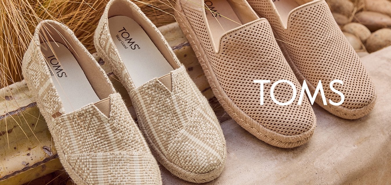 Dva páry béžových nazouvacích bot, jeden s tkanou texturou a druhý s perforacemi, umístěné venku na kamenném povrchu s suchou trávou za nimi.