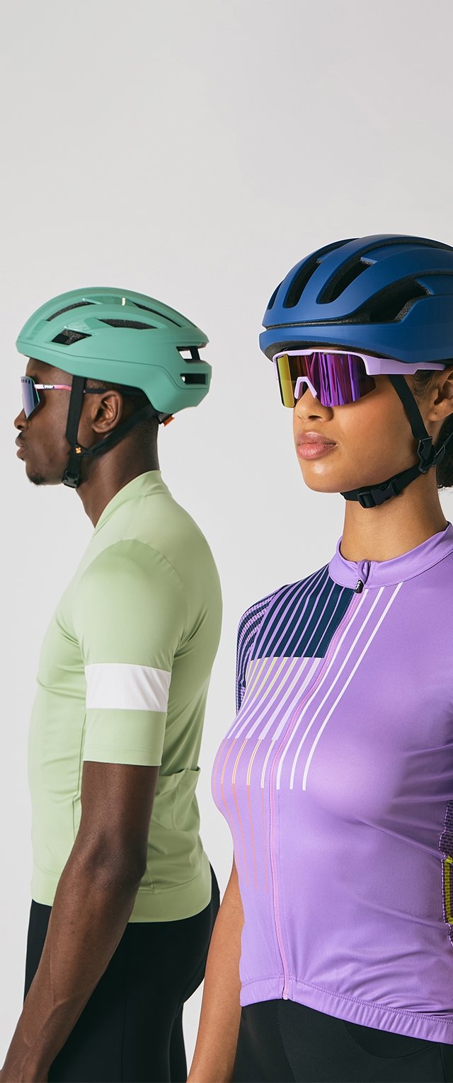 Doi cicliști purtând căști și ochelari de soare reflectorizanți, unul îmbrăcat în tricou ciclist verde și celălalt în tricou ciclist violet, îndreptându-se în direcții opuse.