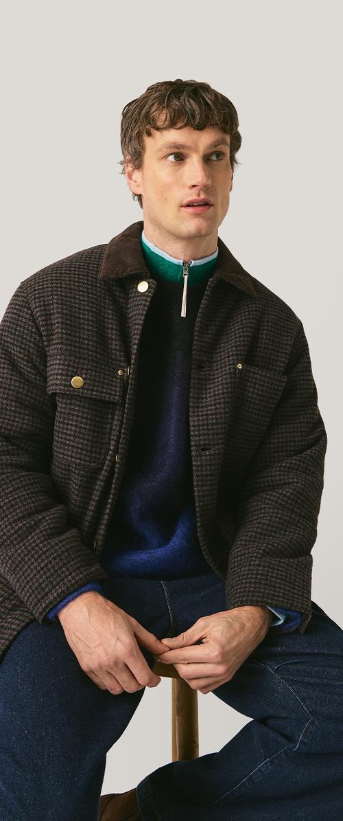 Brun ternet jakke med en mørk krave, to forlommer, båret over en blå sweater. Modellen sidder på en træskammel.