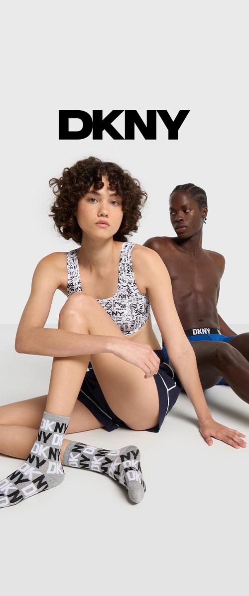 Sujetador deportivo DKNY con diseño gráfico en blanco y negro, pantalones cortos a juego y calcetines grises con logotipo. Dos modelos sentados sobre un fondo neutral.
