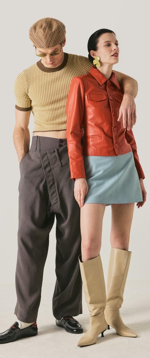 Hombre con camisa beige con estampado, gorro marrón, gafas de sol y pantalones oscuros y holgados con zapatos negros, de pie junto a una mujer con chaqueta de cuero roja, falda azul y botas beige.