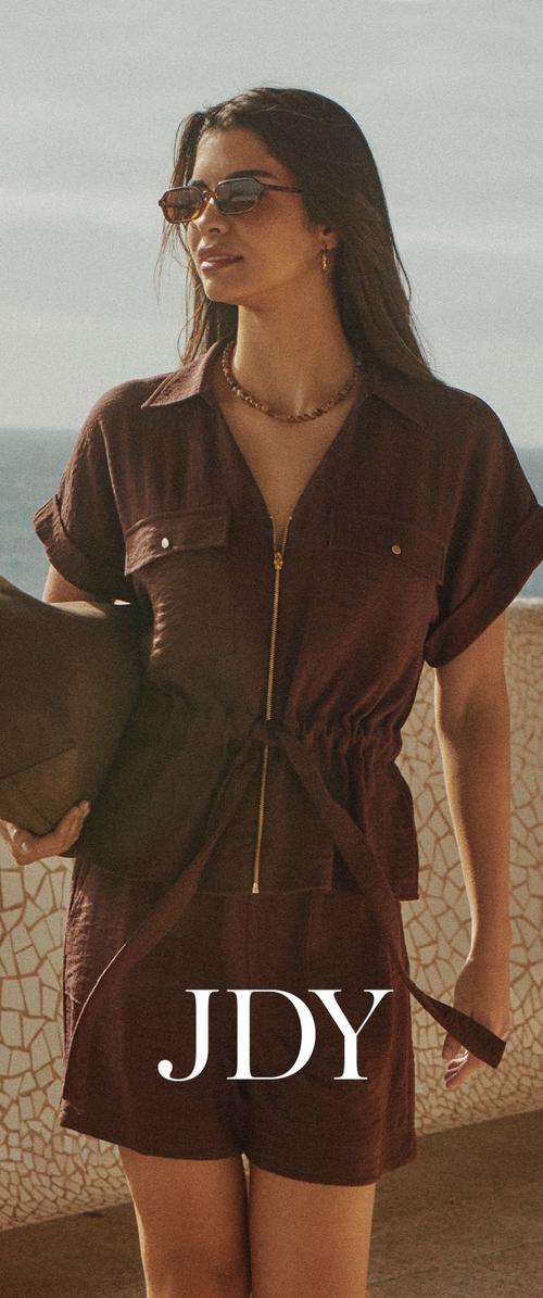 Femme portant une combinaison courte marron avec fermeture éclair, lunettes de soleil, collier, tenant un sac marron, debout près d'un mur de mosaïque avec l'océan en arrière-plan.