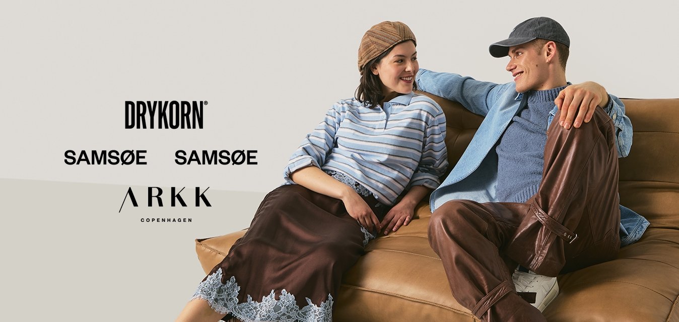 Dva mladí dospělí v běžném oblečení sedí a opírají se o hnědou polstrovanou podlahovou pohovku, usmívají se a dívají se na sebe.