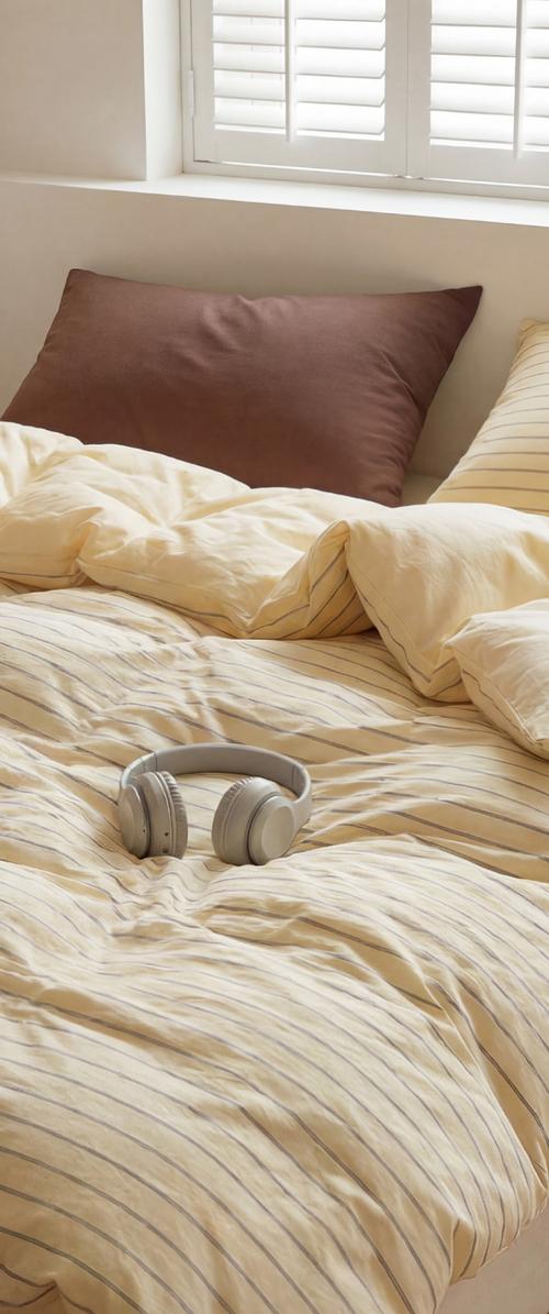 Cojines beige y marrones sobre la cama con ropa de cama crema a rayas y auriculares blancos descansando en la cama cerca de una ventana con contraventanas blancas.