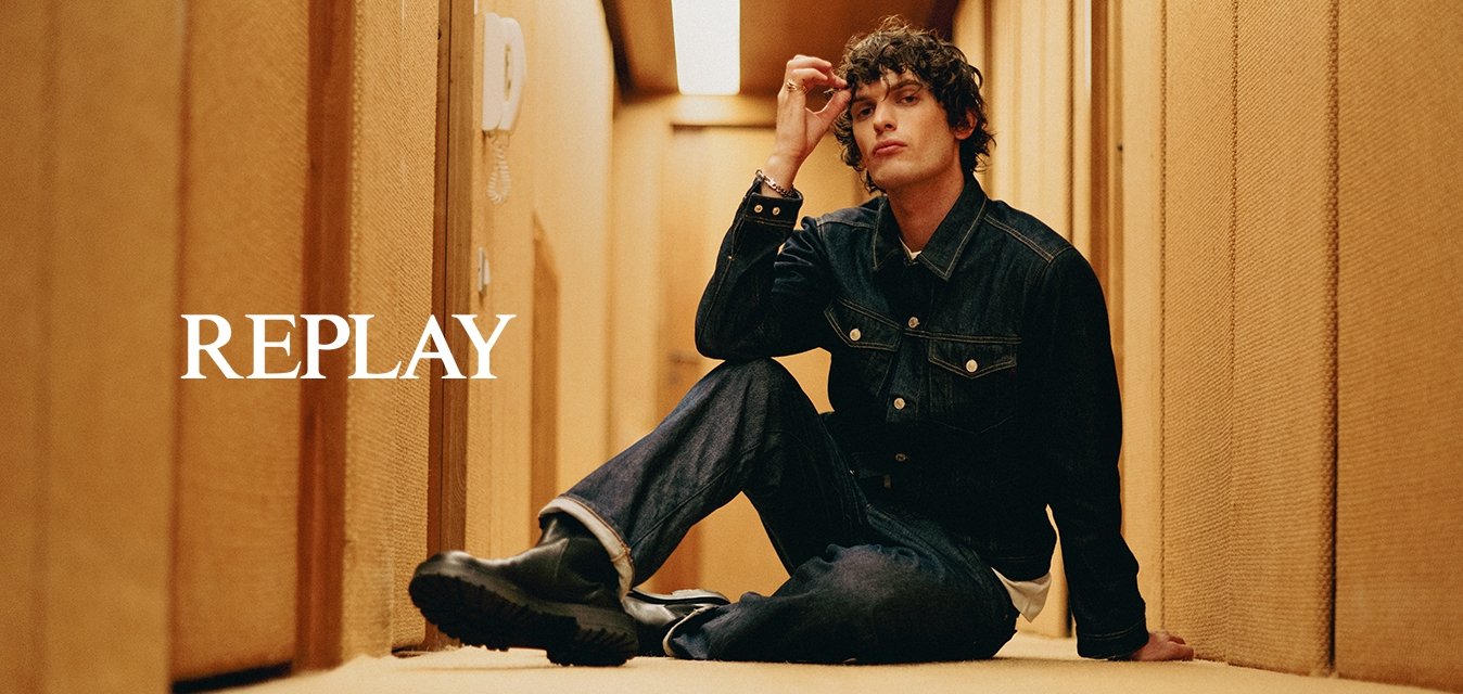 Mladý muž v džínsové bundě a džínách sedí na kobercové podlaze úzké béžové chodby a jednou rukou si dotýká vlasů.