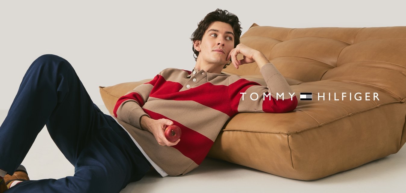 Mladý muž v pruhovaném svetru a modrých kalhotách odpočívá na podlaze, opřený o velké světle hnědé polstrované sezení, drží v ruce červené jablko.