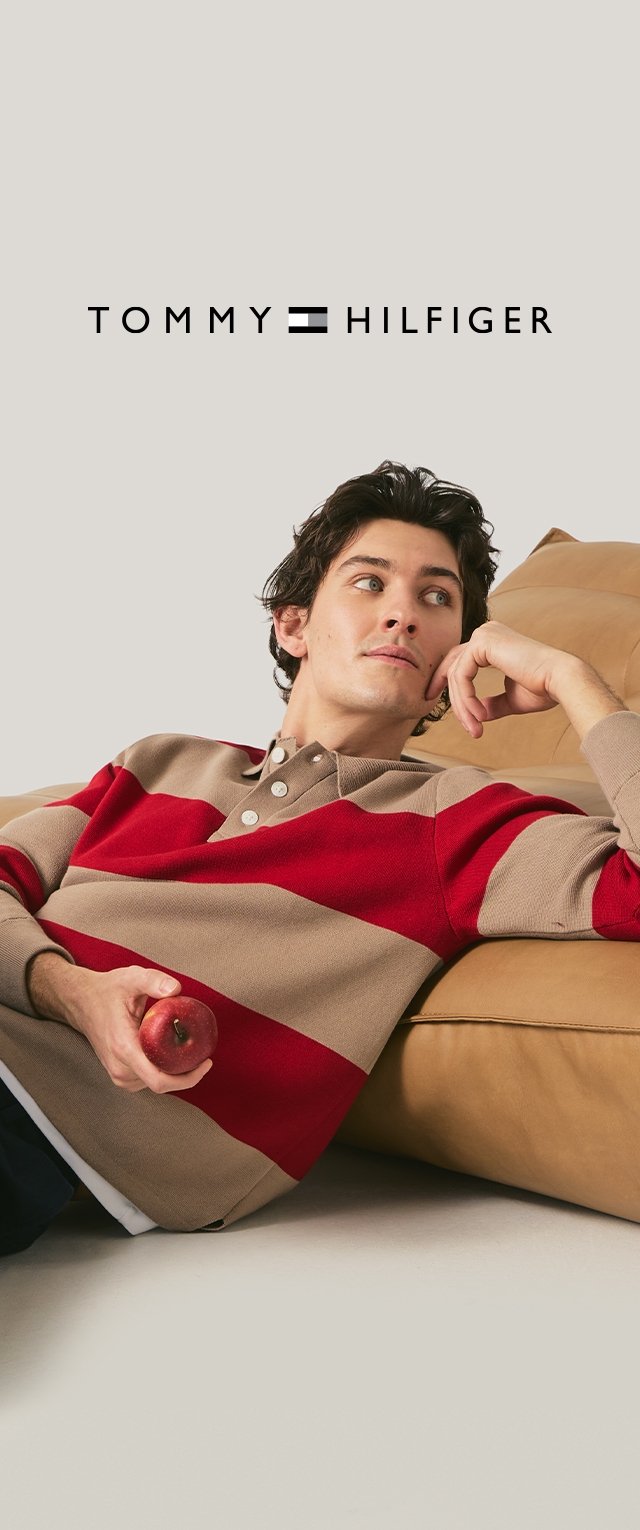 Young man reclining on a tan couch wearing a beige and red striped sweater, holding a red apple with "Tommy Hilfiger" logo above.