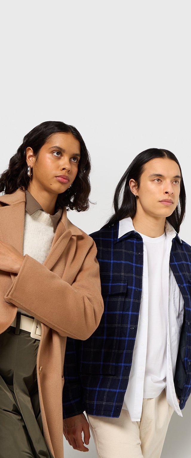 Two models pose together. One wears a long brown coat over a cream knit sweater; the other wears a blue plaid shirt over a white shirt.