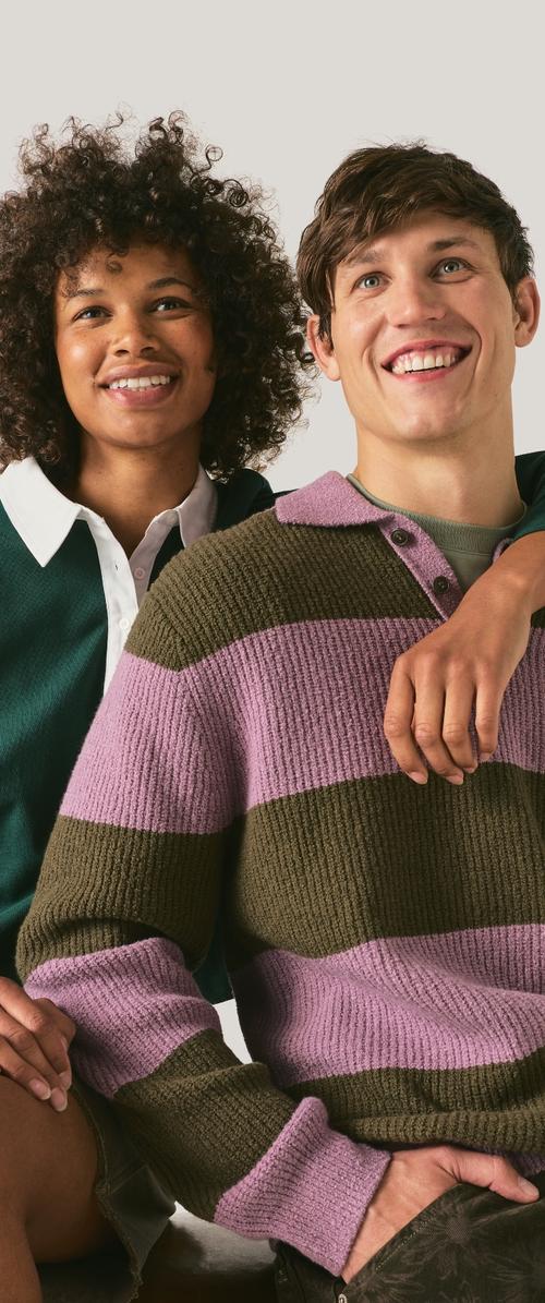 Lachende jonge zwarte vrouw met krullend haar en jonge witte man met kort haar in groen-roze gestreepte trui die dicht bij elkaar zitten, arm om de schouder.