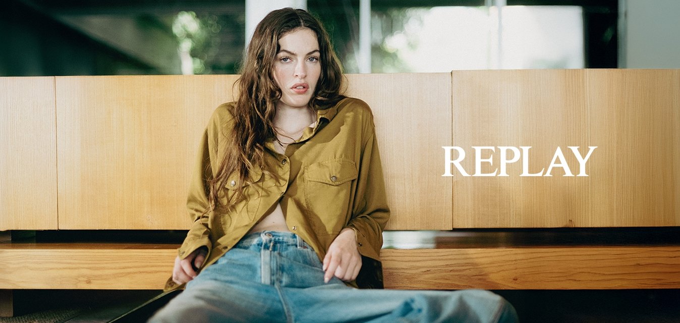 Mladá žena v olivově zelené košili a modrých džínách, sedící na podlaze a opírající se o dřevěnou skříň uvnitř.