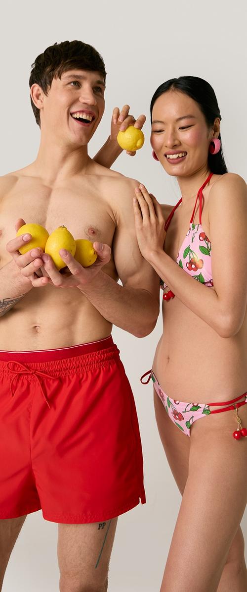 Hombre sonriente sin camiseta con bañador rojo sostiene limones mientras una mujer con bikini floral rosa sostiene juguetonamente un limón cerca de su cabeza.