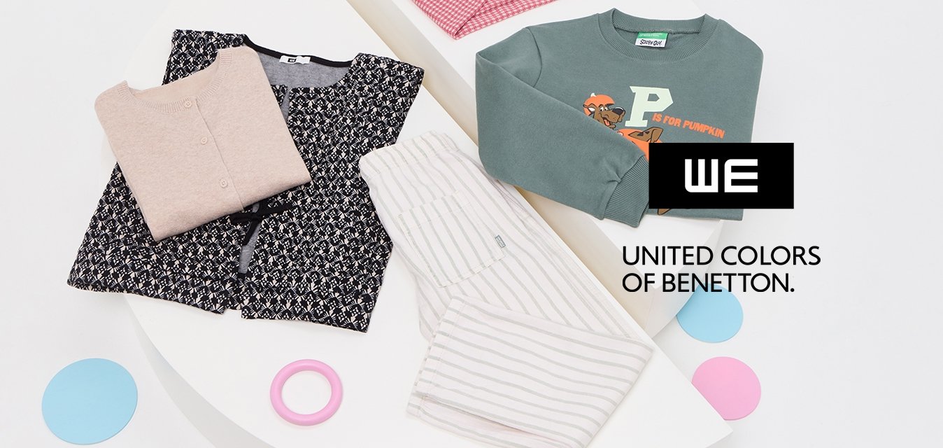 Čtyři složené dětské oděvy—růžový svetr, vzorované černé šaty, pruhované bílé kalhoty a zelená mikina s designem dýně—na bílých stojanech.