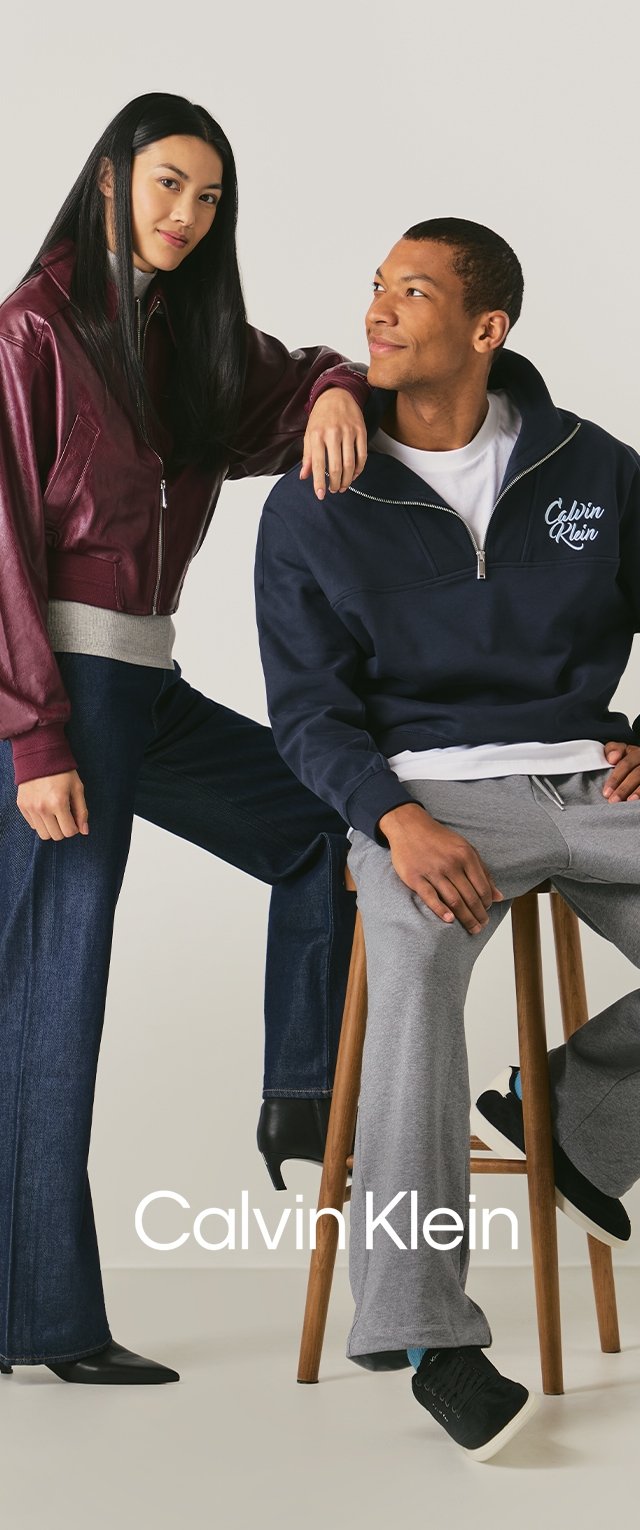 Maroon leather jacket, gray sweater, wide-leg denim pants, navy zip-up hoodie, gray sweatpants, seated on wooden stool, black footwear.