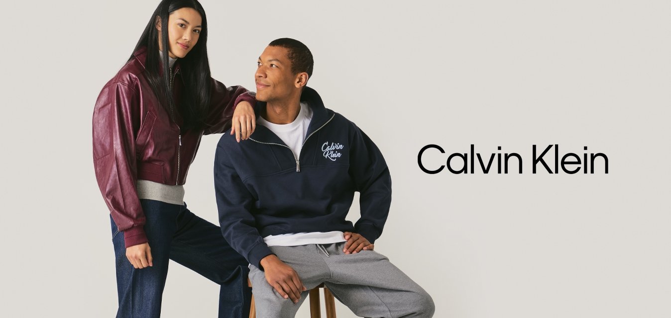 Mladá žena v burgundské bundě a džínách stojí vedle muže v námořnické mikině a šedých kalhotách, který sedí na stoličce proti jednoduchému pozadí.