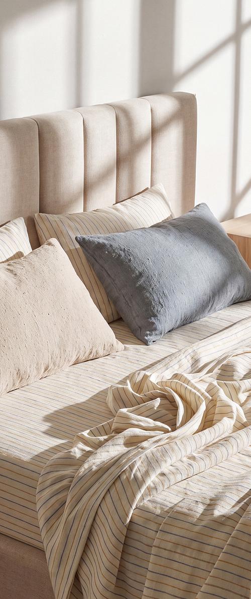 Beige polstret seng med stripete beige og lyseblå sengetøy, beige og grå puter, og sollys som kaster skygger på hodegjerde.