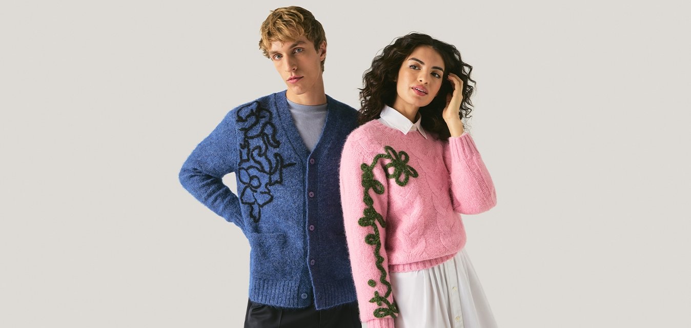 Muž v modrém cardiganu s černým květinovým vzorem a žena v růžovém svetru se zeleným květinovým vzorem a bílou sukní stojí na světlém pozadí.