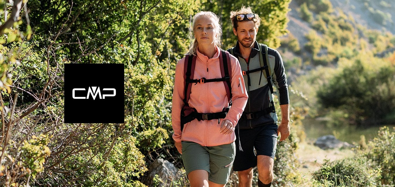 Dva turisté v outdoorovém oblečení jdou po prašné cestě vedle řeky, obklopeni zelenými stromy a kopci na slunci.