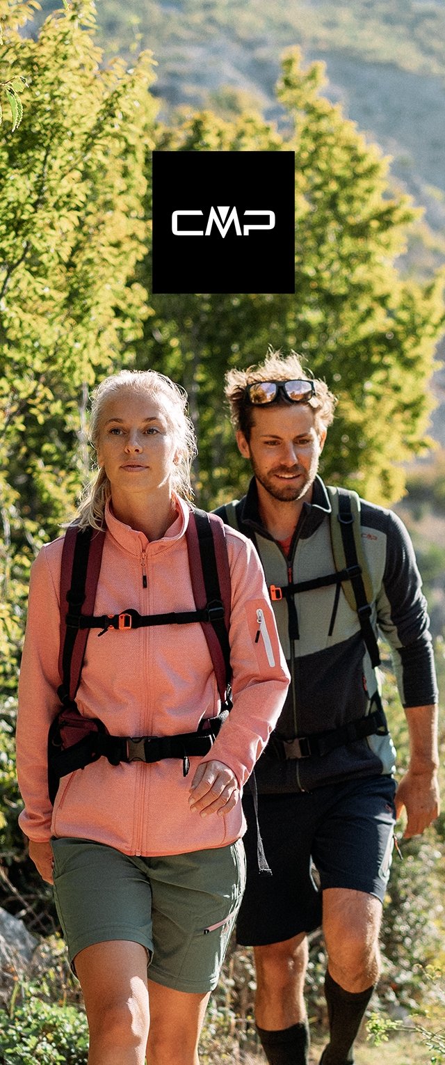 Dos excursionistas con mochilas y ropa de exterior caminando por un sendero forestal iluminado por el sol con follaje verde alrededor.