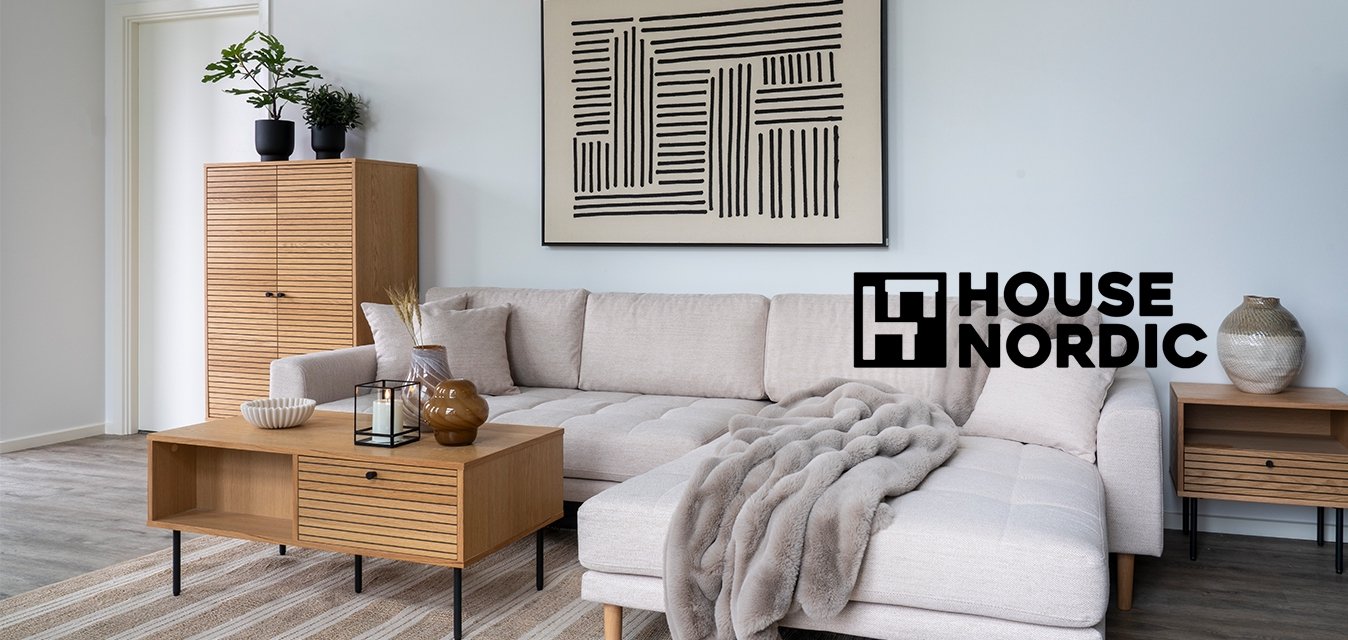 Béžová látková rohová pohovka na dřevěných nohou, umístěná vedle pruhovaného koberce. Obsahuje dřevěný konferenční stolek se zásuvkami a doplňky.