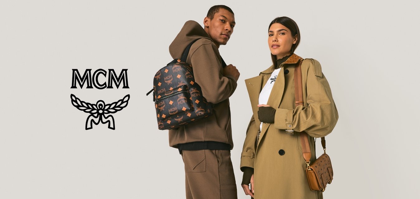 Muž v hnědé mikině s kapucí a kalhotách s barevným vzorovaným batohem; žena v dlouhém béžovém kabátu a bílém topu držící hnědou kabelku přes rameno, stojící.