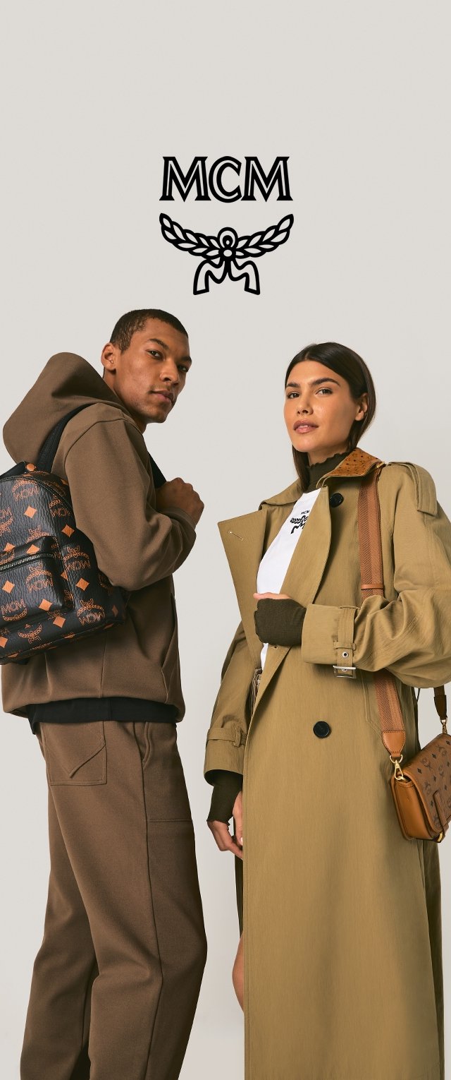 Dwóch modeli pozujących z produktami MCM. Mężczyzna ma na sobie brązową bluzę z kapturem i spodnie, trzymając czarną torbę w wzory. Kobieta nosi beżowy trencz, białą koszulę i niesie brązową torebkę z ozdobnymi akcentami.