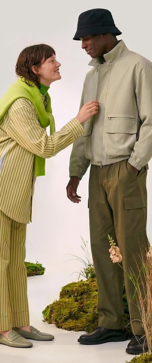 A smiling woman adjusts a man's beige jacket zipper, both wearing casual green and earth-toned outfits, standing on moss with flowers nearby.