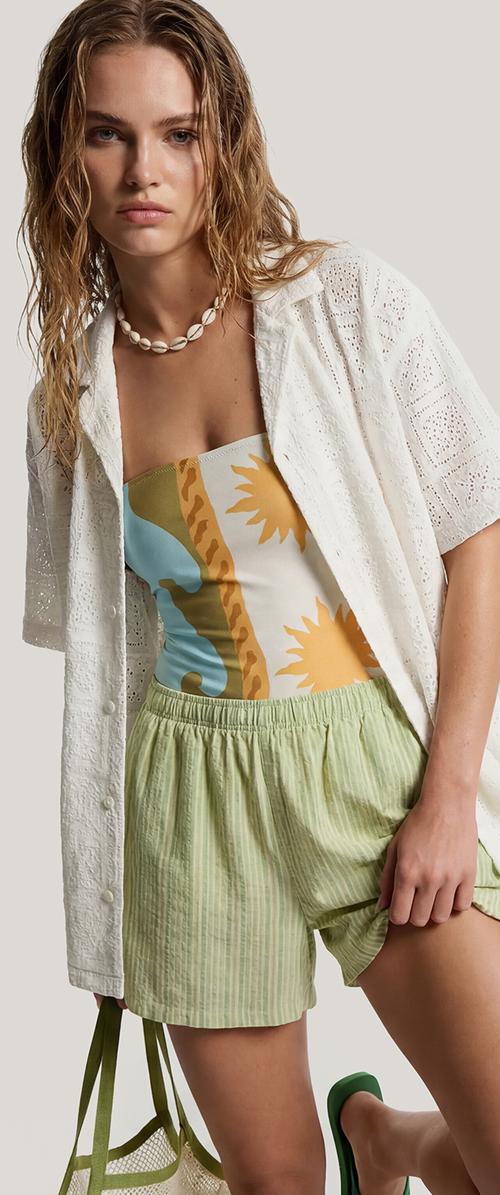 Jeune femme aux cheveux mouillés porte une chemise blanche en broderie anglaise, un top tube à motif, un short vert clair, un collier de coquillages et tient un sac en filet.