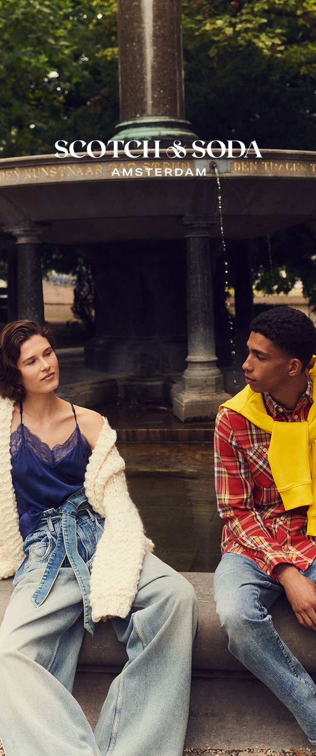 Due modelli sono seduti vicino a una fontana. Uno indossa un cardigan a coste color crema su una maglietta blu e pantaloni di jeans larghi. L'altro indossa una camicia a quadri e un maglione giallo.