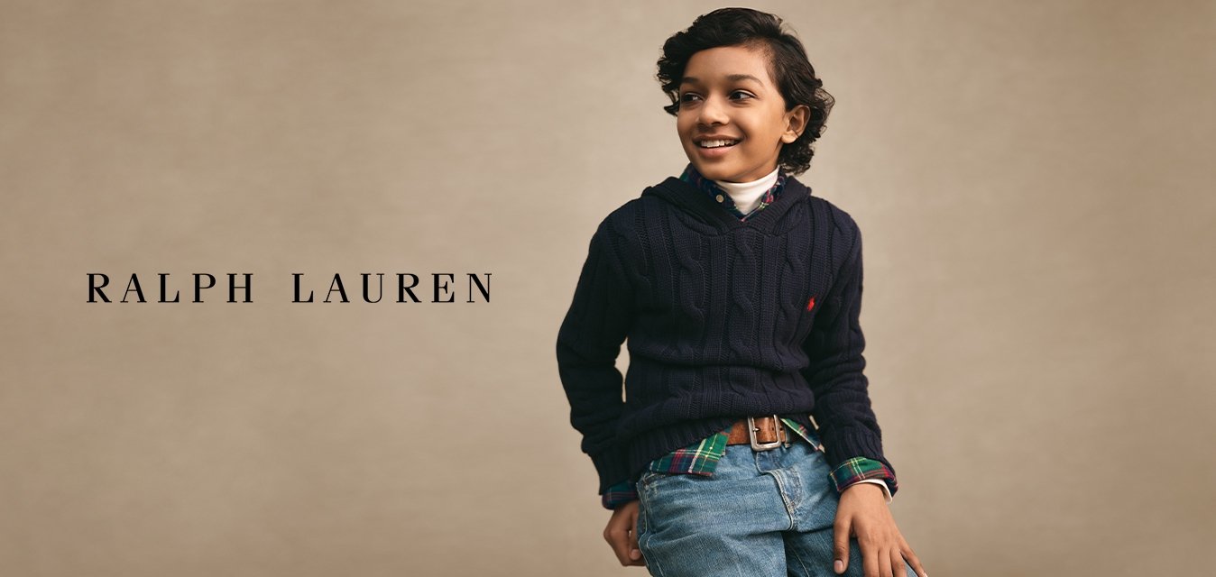 Chlapec s tmavými kudrnatými vlasy, oblečený v tmavě modrém svetru přes kostkovanou košili a džíny, usmívající se a dívající se stranou na jednobarevném pozadí.