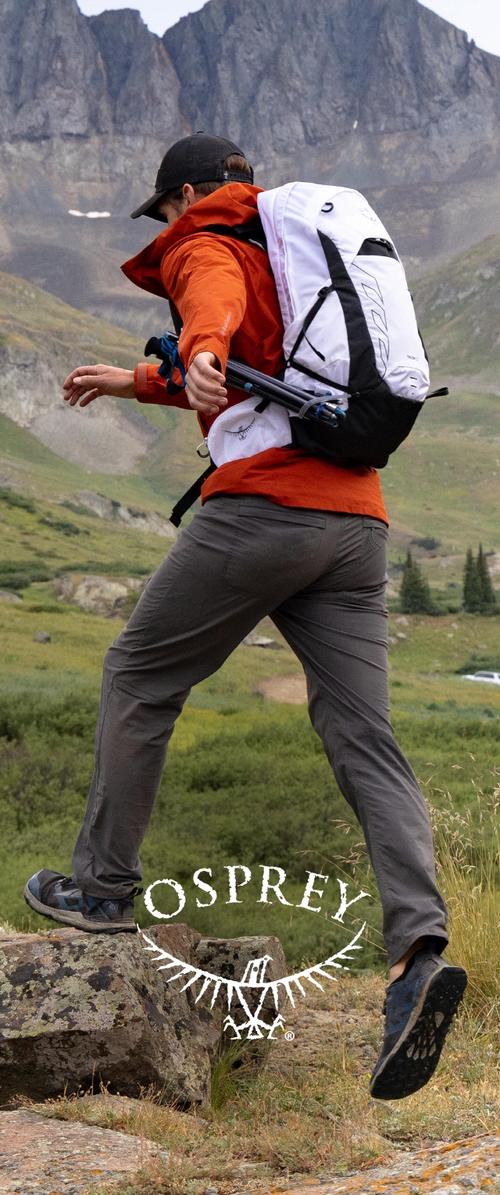 Man in orange jacket and gray pants steps over a rock, carrying a white Osprey backpack with blue hardware and adjustable straps.