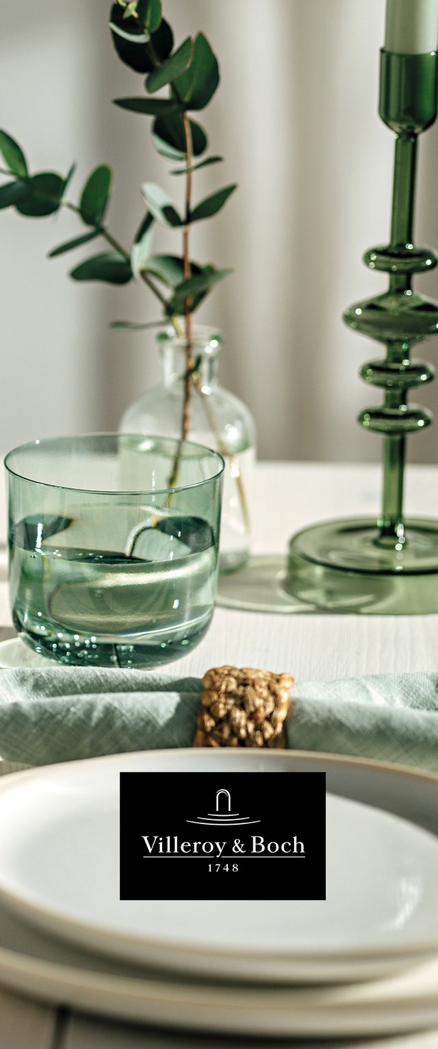 Platos blancos apilados con una servilleta verde doblada sujeta por un anillo tejida, junto a un vaso de agua, una planta en un jarrón transparente y un portavelas verde.