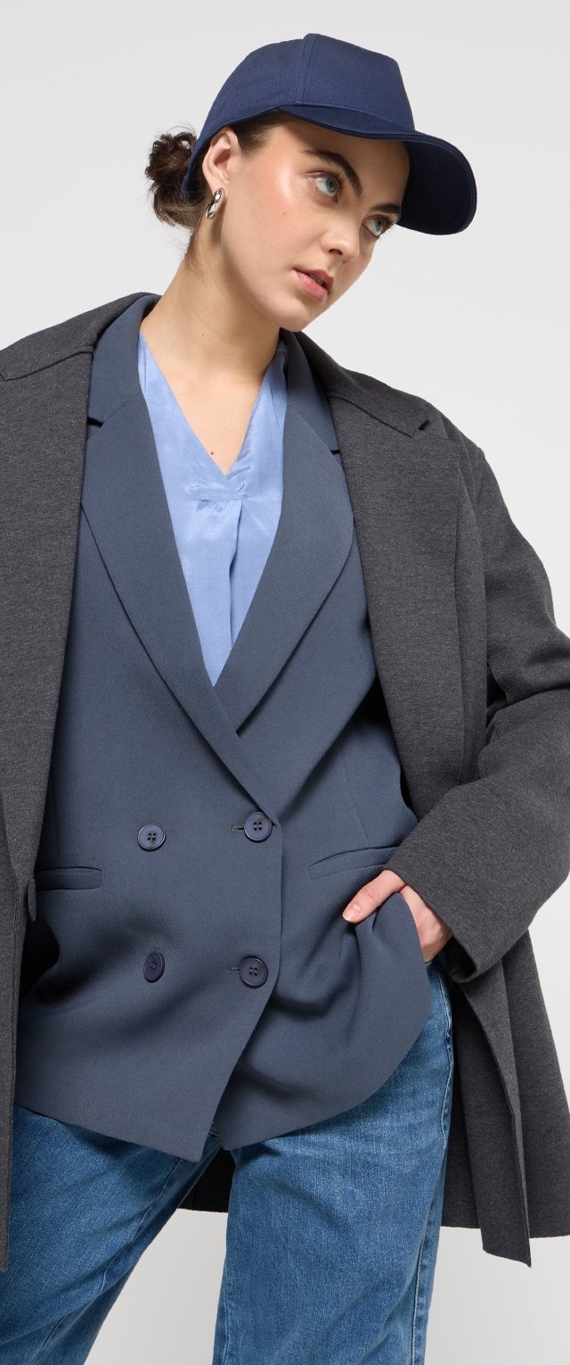 Grauem Mantel über einem blauen, zweireihigen Blazer, kombiniert mit einer hellblauen Bluse und Jeans. Das Outfit umfasst eine marineblaue Mütze und auffällige Ohrringe.
