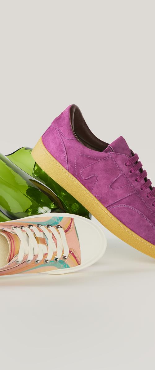 Sneaker in camoscio viola con suola in gomma appoggiata a una sneaker in tela multicolore con lacci bianchi, accanto a una bottiglia di vetro verde su uno sfondo chiaro.