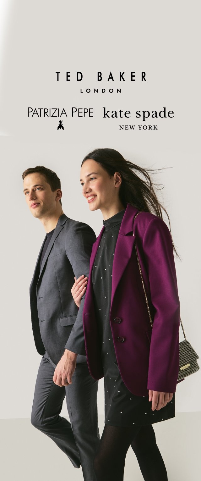 Zwei Models gehen nebeneinander her. Der Mann trägt einen dunkelgrauen Anzug, und die Frau trägt ein lila Blazer über einem schwarzen Kleid mit weißen Punkten.