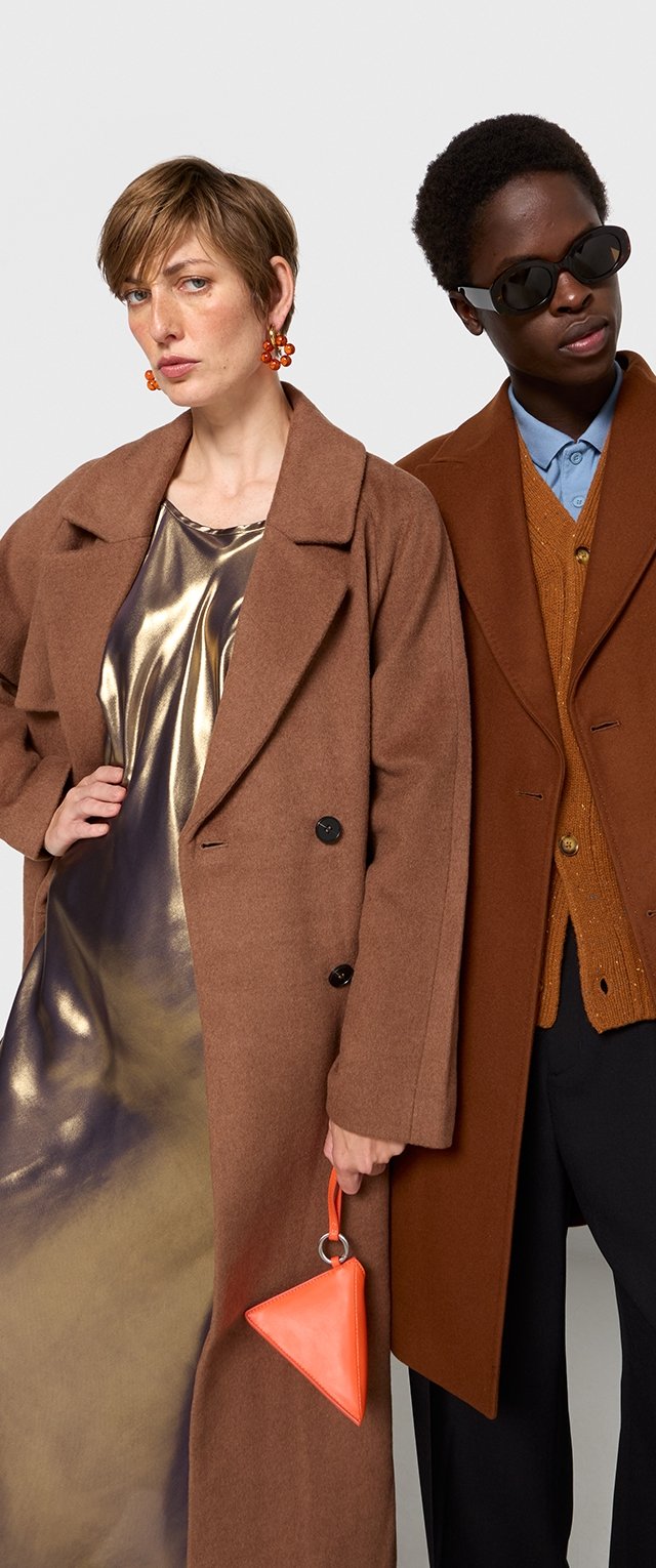 Dos modelos con ropa de abrigo: uno lleva un abrigo de lana marrón, y el otro un abrigo marrón con un suéter naranja texturizado. Los accesorios incluyen un bolso triangular naranja.