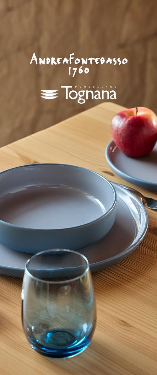 Blaues Porzellanteller-Set auf einem Holztisch, bestehend aus einem großen runden Teller, einem kleineren Teller, einem Glas mit blauem Boden und einem roten Apfel.